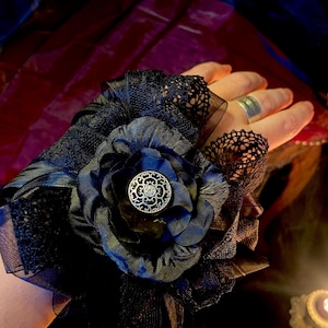 Op de afbeelding: Zwarte vingerloze handschoen met een grote, donkerblauwe stoffen bloem en een decoratieve zilveren knoop. De handschoen heeft lagen zwart kant en tule, wat een gotische of Victoriaanse esthetiek creëert.