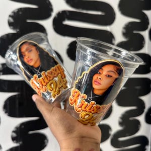 May include: Two clear plastic cups featuring a custom graphic print of a person with long dark hair wearing a silver heart necklace. Below the portrait, the text Krissy 24 is displayed in a bold, orange, and yellow gradient font.