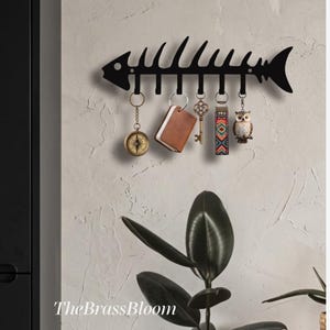 May include: A black metal wall-mounted key holder shaped like a fish skeleton with six hooks. Hanging from the hooks are a round compass keychain, a brown rectangular leather-like tag, an ornate brass-coloured key, a colourful patterned fob, and an owl.