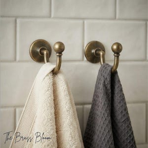 May include: Two antique brass wall hooks with a rounded ball finial design. A cream-coloured herringbone towel hangs from the left hook, while a dark grey waffle-textured towel hangs from the right hook. The text The Brass Bloom is visible.