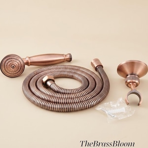May include: A copper-toned handheld shower head with a matching flexible metal hose and a wall-mounted bracket. The set includes a small plastic bag containing mounting hardware. The text TheBrassBloom is visible in the bottom right corner.