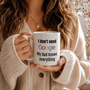 May include: White ceramic mug with the text "I don't need Google, My Dad Knows everything" in black and colorful lettering. The mug is held by a person wearing a beige sweater.