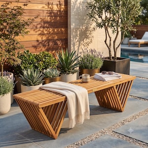 May include: A slatted teak wood bench with triangular side supports, featuring a cream-colored fringed throw blanket draped over the seat. A ceramic mug and a stack of magazines rest on the surface, with a small metal tag labeled TURA.