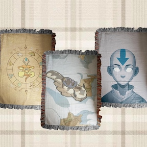 May include: Three rectangular woven throw blankets with fringed edges. The left blanket is tan with a circular emblem featuring a central figure. The centre blanket is light blue with a white creature. The right blanket is grey with a portrait of a bald figure.