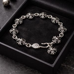 May include: A silver bracelet with a floral design, presented in a black box. The bracelet features multiple flower-shaped links connected by small rings. The clasp and a charm also have a flower design. Two silver spheres are visible in the background.