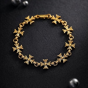May include: A gold bracelet featuring a repeating floral cross pattern. The bracelet is composed of linked segments, each with a stylized cross design. The clasp is visible. The bracelet is set against a dark background.