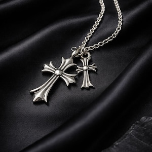 Könnte beinhalten: Eine silberfarbene Halskette mit Anhänger, die zwei unterschiedlich große Kreuzanhänger zeigt. Das größere Kreuz hat ein detailliertes, verziertes Design. Die Halskette und Kreuze sind auf schwarzem, strukturiertem Untergrund dargestellt.