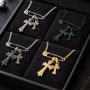 May include: A black jewelry box displaying four cross pendant necklaces. The necklaces are silver, black, and gold, each attached to a chain with a safety pin clasp. The crosses have decorative details.