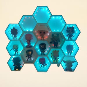 Puede incluir: Una estantería hexagonal con retroiluminación LED azul, que muestra varias figuras de personajes. La estantería está compuesta por múltiples compartimentos interconectados en forma de panal, cada uno con una figura diferente. El diseño es moderno y atractivo.