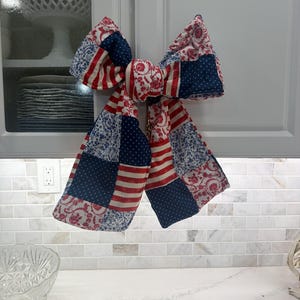 May include: A decorative fabric bow featuring a patchwork design with red, white, and blue colors. The pattern includes sections of red and white stripes, blue fabric with white polka dots, and white fabric with a red floral print motif.