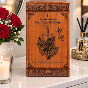 Puede incluir: Una urna rectangular de madera con acabado marrón medio, que presenta un grabado de manos en oración, una espada, rosas y dos pájaros. El texto dice: Be Not Afraid, For I Am With You, ISAIAH 41:10, y una cita inspiradora más larga.