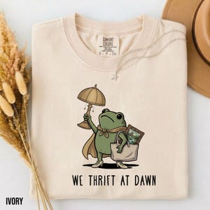 May include: An ivory t-shirt featuring a graphic of a green frog wearing a cape, holding a lamp, and carrying a tote bag with a framed picture inside. The text below the frog reads WE THRIFT AT DAWN. The shirt has a crew neckline and short sleeves.