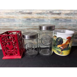May include: A red metal utensil holder, two glass jars with silver lids and rooster emblems, and a ceramic utensil holder with a rooster design. The items are arranged on a black surface against a wood-paneled background.