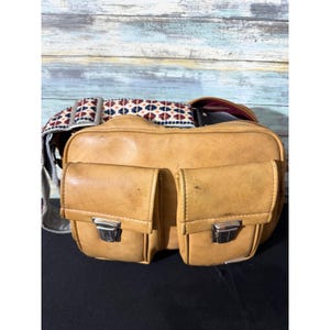 Puede incluir: Un bolso bandolera de cuero color canela con dos bolsillos frontales y cierres de presión de metal plateado. El bolso incluye una correa de hombro ancha y ajustable decorada con un patrón geométrico repetitivo en rojo, azul y blanco.