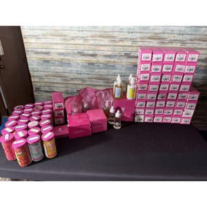 May include: A collection of beauty products, including pink boxes, clear bottles, and jars filled with colorful sprinkles. The items are arranged on a black surface against a wooden background. The products appear to be ready for sale.