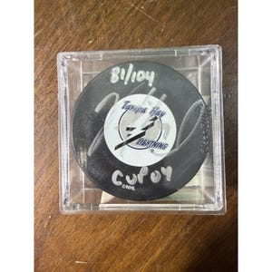 May include: A black Tampa Bay Lightning hockey puck encased in a clear square plastic display box. The puck features a white center logo, a silver signature, the handwritten text 81/104 and Cup 04, and the NHL logo printed at the bottom edge.