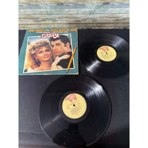 May include: A vintage vinyl record album of the movie "Grease" featuring John Travolta and Olivia Newton-John. The album cover is light blue with a photo of the stars. Two black vinyl records are also visible.