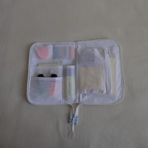 May include: A white zippered organizer case featuring multiple translucent mesh pockets. The interior contains various stationery items, including a pink circular sticky note, colorful rectangular page markers, a highlighter, and pens.