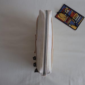 May include: A cream-colored canvas pencil case with a silver metal zipper closure. The side of the case features three small black pens with visible nib sizes clipped to the exterior. A rectangular sticker with the word STANDOUT is nearby.