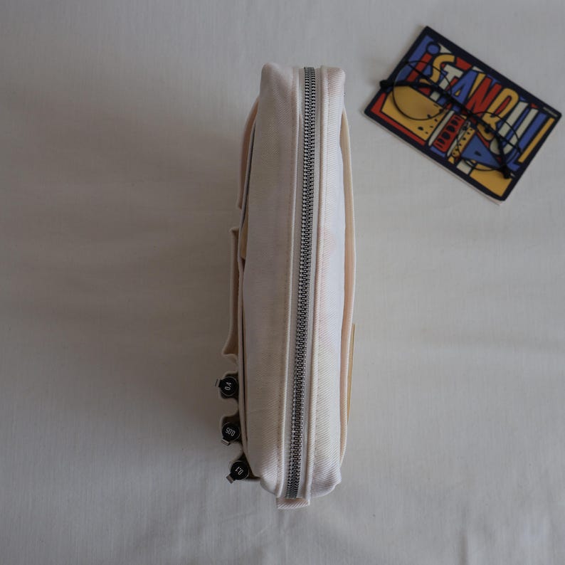 May include: A cream-colored canvas pencil case with a silver metal zipper closure. The side of the case features three small black pens with visible nib sizes clipped to the exterior. A rectangular sticker with the word STANDOUT is nearby.
