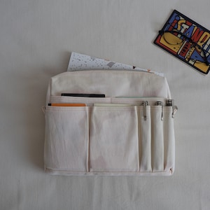 May include: A cream-colored canvas bag organizer featuring a zippered main compartment and multiple exterior pockets of varying sizes. The front includes several vertical slots for pens or pencils and wider pockets holding small notebooks and papers.