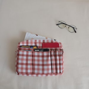 May include: A red and white gingham patterned organizer with multiple pockets, holding cards, a red notepad, and pens. A pair of round-framed glasses rests nearby. The organizer is designed for desk or bedside use.