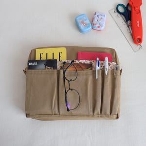 May include: A beige organiser bag with several pockets, containing a yellow magazine, a red notebook, glasses, pens, and a card. Two small, decorated containers, scissors, a ruler, and a red cutter are also visible.
