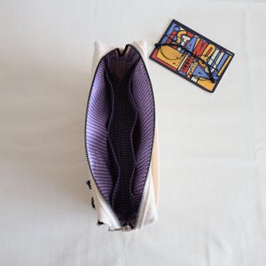 May include: An open off-white pouch with a purple and white gingham interior lining featuring multiple internal compartments. Beside the pouch lies a rectangular card with the word ISTANBUL printed in bold letters and a pair of thin black glasses.