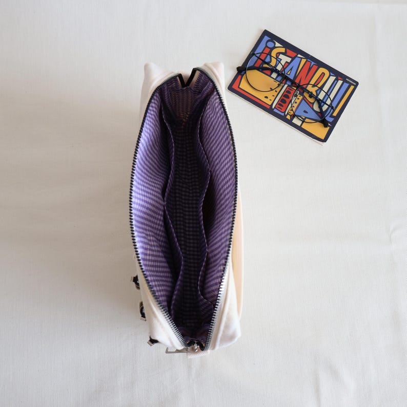May include: An open off-white pouch with a purple and white gingham interior lining featuring multiple internal compartments. Beside the pouch lies a rectangular card with the word ISTANBUL printed in bold letters and a pair of thin black glasses.