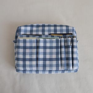 May include: A blue and white gingham fabric organizer with multiple pockets. The organizer is rectangular and has several vertical pockets on the front. Pens and papers are visible in the pockets. The fabric is a classic checkered pattern.