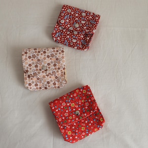 May include: Three small, square fabric pouches with floral patterns. One is red with white and blue flowers, another is a light peach with brown and white flowers, and the third is red with yellow and blue flowers. Each pouch has a silver snap closure.