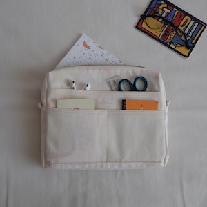May include: A cream-colored canvas organizer pouch featuring multiple exterior pockets. The pockets contain white wireless earbuds, a small clear ruler, teal-handled scissors, a cream notebook, and an orange notepad. A zipper runs along the top.