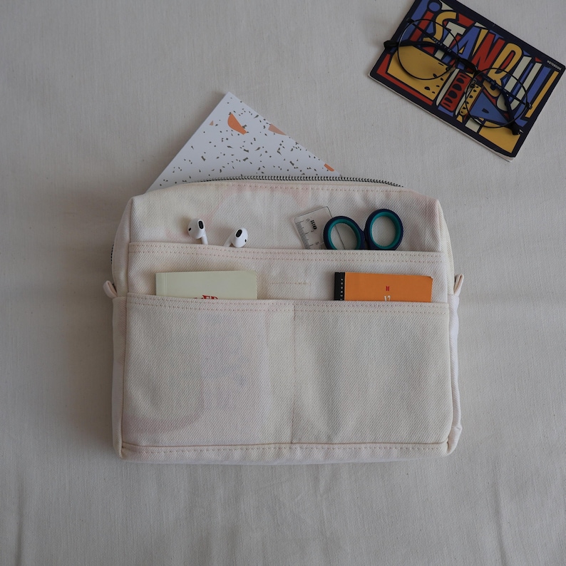 May include: A cream-colored canvas organizer pouch featuring multiple exterior pockets. The pockets contain white wireless earbuds, a small clear ruler, teal-handled scissors, a cream notebook, and an orange notepad. A zipper runs along the top.