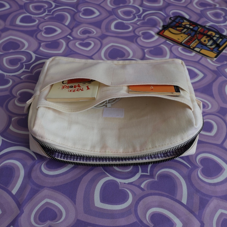 May include: A cream-colored canvas organizer pouch with a zippered main compartment and two exterior slip pockets. One pocket contains a small book with the text I NEED MORE, while the other holds a thin orange notebook and a white charging cable.