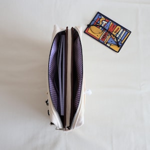 May include: An open cream-colored canvas pencil case with a purple and white gingham interior lining. The case contains a notebook and is positioned next to a rectangular card featuring the word STANDOUT and a pair of black-rimmed glasses.