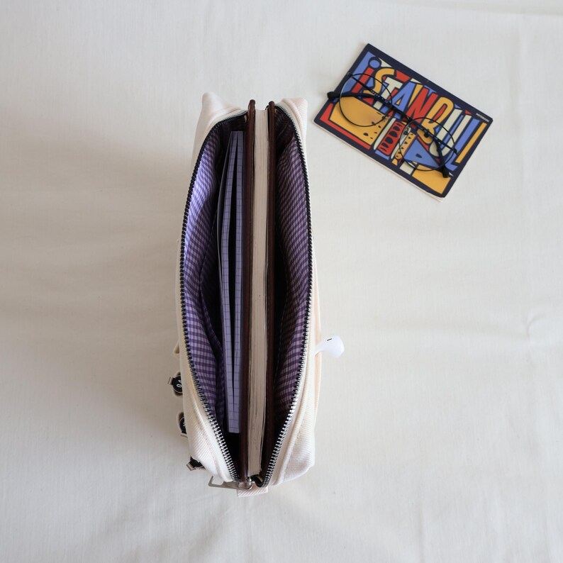 May include: An open cream-colored canvas pencil case with a purple and white gingham interior lining. The case contains a notebook and is positioned next to a rectangular card featuring the word STANDOUT and a pair of black-rimmed glasses.