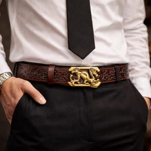 Op de afbeelding: Bruine leren riem met een reliëf bloemmotief en een goudkleurige draakgesp. De riem wordt gedragen met een wit overhemd, een zwarte stropdas en een zwarte broek. De gesp is rechthoekig en heeft een gedetailleerd drakenontwerp.