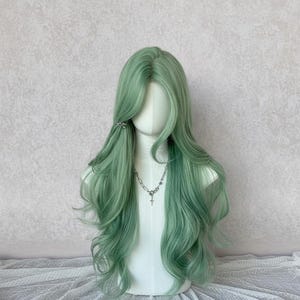 May include: A long, wavy wig in a soft, muted green hue, styled on a mannequin head. The wig features a side part, with long layers and a few strands pinned back with a small silver clip. A silver necklace with a cross pendant adds a subtle detail.