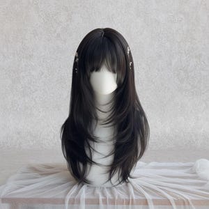 Könnte beinhalten: Schwarze Perücke mit langem, gestuftem Haar und Pony, auf einem Schaufensterkopf präsentiert. Die Perücke hat einen natürlich aussehenden Haaransatz und ist mit kleinen, dekorativen Haaraccessoires verziert. Das Haar fällt über die Schultern.