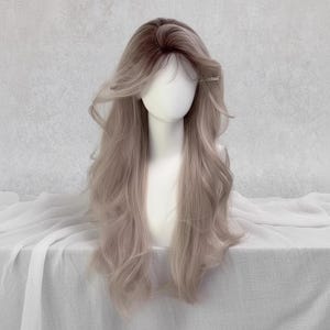 May include: A long, wavy wig with a dark brown to light brown ombre effect. The wig is styled with a side part and soft layers, and is displayed on a mannequin head. The hair has a natural-looking shine and is held in place with a small hair clip.