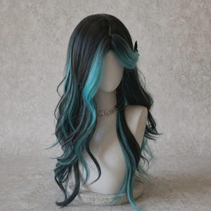 Pode incluir: Uma peruca longa e ondulada com uma base de cabelo preto profundo e madeixas vibrantes em turquesa que emolduram o rosto e caem ao longo do comprimento. O cabelo tem uma risca ao meio e está num manequim com um colar de corrente prateada.