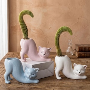 Puede incluir: Tres macetas de cerámica con forma de gato en colores pastel: azul claro, rosa y blanco. Cada maceta presenta un diseño en forma de gato con un cuerpo curvado y una planta de cactus. Los gatos están en posición de estiramiento, con los ojos cerrados.