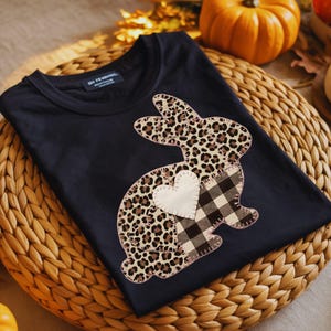 Puede incluir: Camiseta negra con un diseño de conejito. El conejito presenta un estampado de leopardo y un parche en forma de corazón, con un patrón de cuadros en la parte inferior del cuerpo. La camiseta está doblada sobre una superficie tejida, con calabazas y hojas en el fondo.