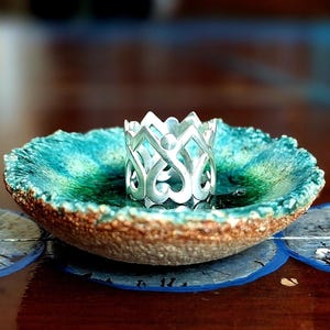 Puede incluir: Un cuenco decorativo de cerámica turquesa y marrón con un portavelas de metal plateado. El cuenco está colocado sobre una superficie de madera oscura con diseños de media luna azules.