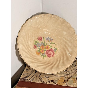 Könnte beinhalten: Ein cremefarbener, dekorativer Vintage-Teller mit gewelltem Rand. Der Teller hat ein zentrales Kreuzstich-Blumenmuster in Rosa, Blau, Gelb und Grün. Der Teller hat ein Wirbelmuster und einen goldenen Rand.
