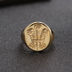 May include: A gold-toned signet ring featuring an engraved Prince of Wales feathers emblem on the circular face. The design includes three ostrich feathers emerging from a coronet with a ribbon banner below displaying the text ICH DIEN.