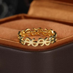 Puede incluir: Un anillo dorado con un diseño repetido de signos de dólar. El anillo se muestra sobre un fondo marrón, posiblemente dentro de una caja de joyería. El diseño del anillo es abierto, permitiendo que la luz pase a través de las formas de signos de dólar.