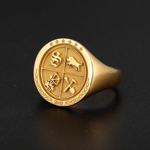 May include: A gold-toned signet ring featuring a circular face divided into four quadrants. The quadrants display an ornate letter S, a bull, an archer, and crossed tools. The border includes five stars at the top and a laurel leaf pattern.