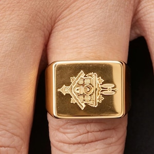May include: A gold-toned signet ring featuring a rectangular face with a detailed, laser-engraved illustration of a traditional cuckoo clock. The engraving depicts the clock's house-like structure, a central dial, and hanging weights on the side.