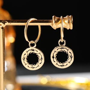 May include: Gold-colored hoop earrings with circular pendants. Each pendant features a stylized, swirling design around a central opening. The earrings are displayed on a gold-toned jewelry stand against a dark background.
